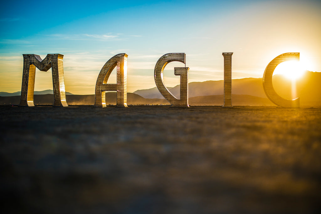 Large metallic letters spelling MAGIC are arranged outdoors in a desert landscape at sunset, with mountains in the background and the sun shining behind the final letter.