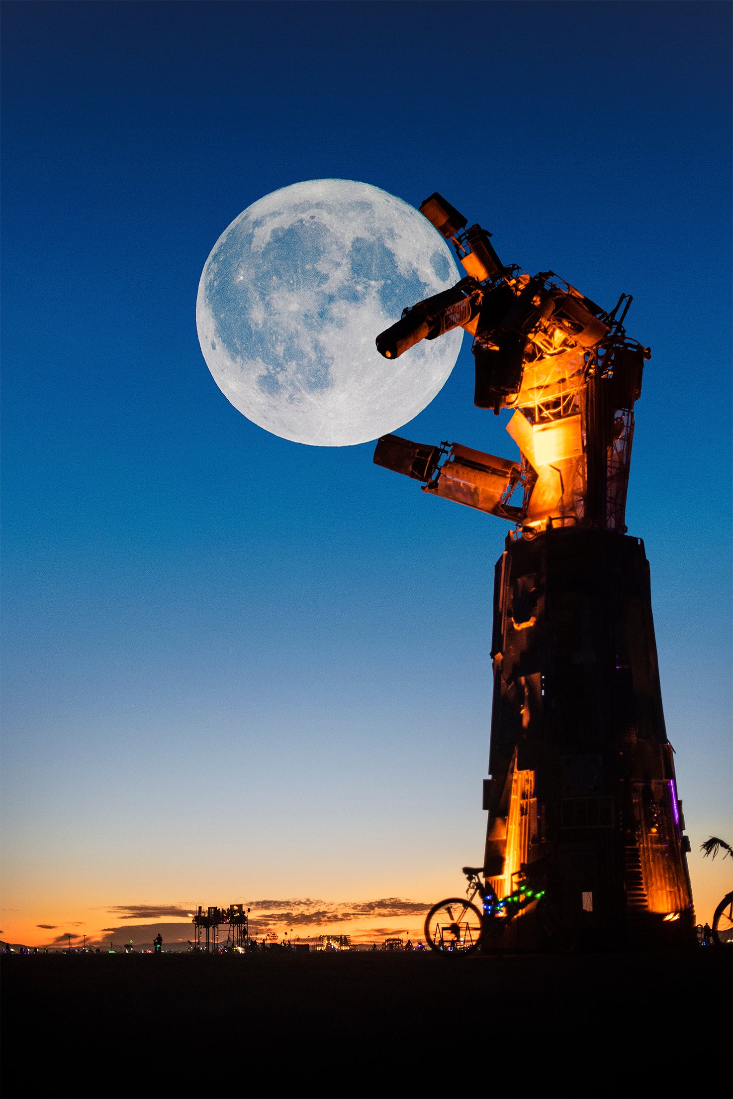 A large illuminated moon sculpture at dusk, with a giant full moon in the blue twilight sky above a silhouetted landscape and small distant lights. burningman photography