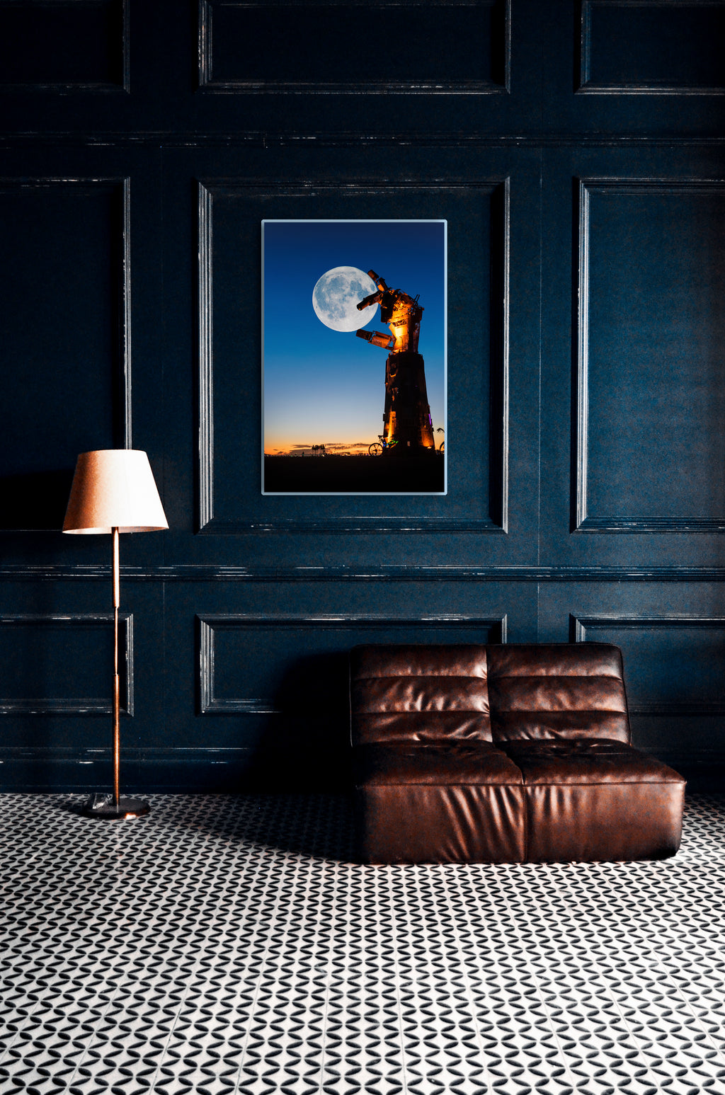 A framed vertical art print depicting a silhouetted figure holding a large moon-like sphere against a blue sky, displayed on a dark navy paneled wall above a brown leather sofa with a patterned black-and-white floor and a floor lamp. burning man photo
